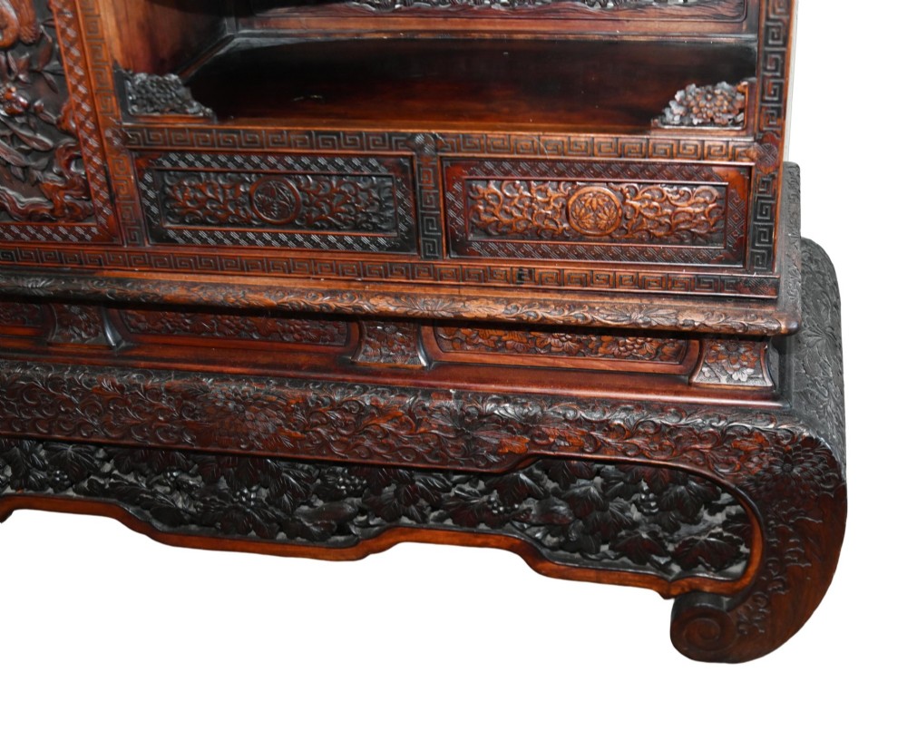 An intricately carved dark brown Japanese Meiji period display cabinet featuring asymmetrical open shelves, relief-carved cupboard doors, and a decorative cresting piece. View VI