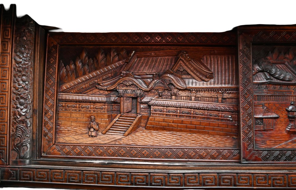 An intricately carved dark brown Japanese Meiji period display cabinet featuring asymmetrical open shelves, relief-carved cupboard doors, and a decorative cresting piece. View IX