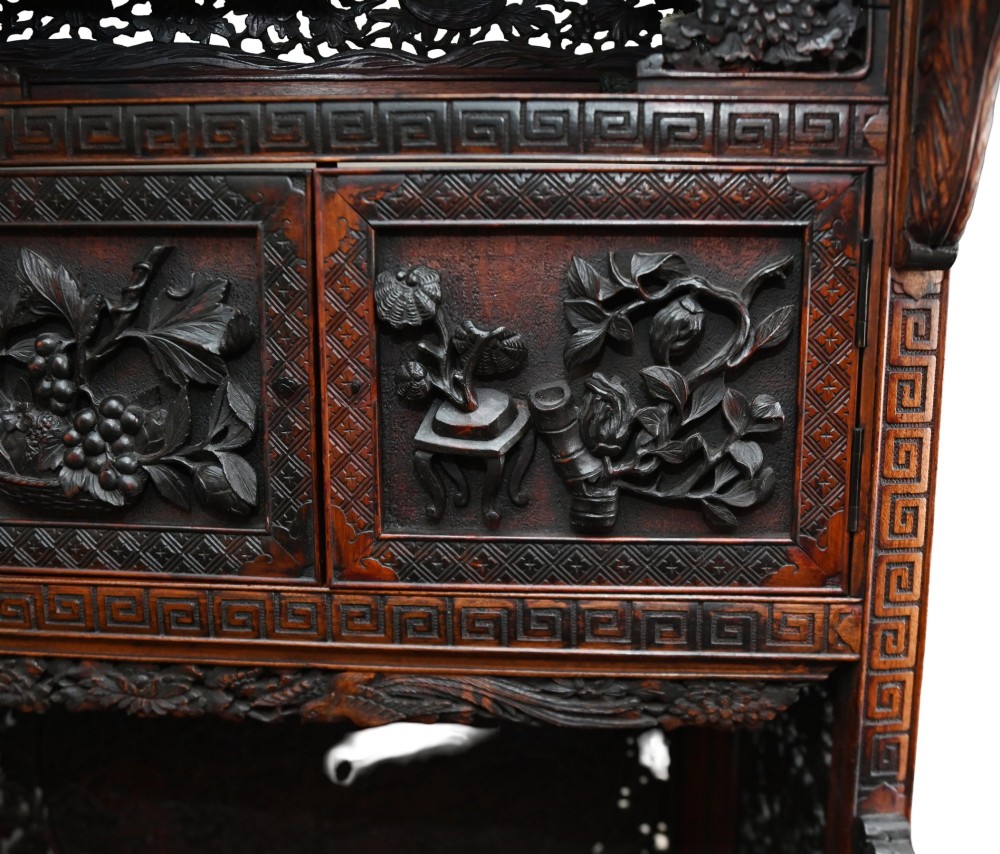 An intricately carved dark brown Japanese Meiji period display cabinet featuring asymmetrical open shelves, relief-carved cupboard doors, and a decorative cresting piece. View XI