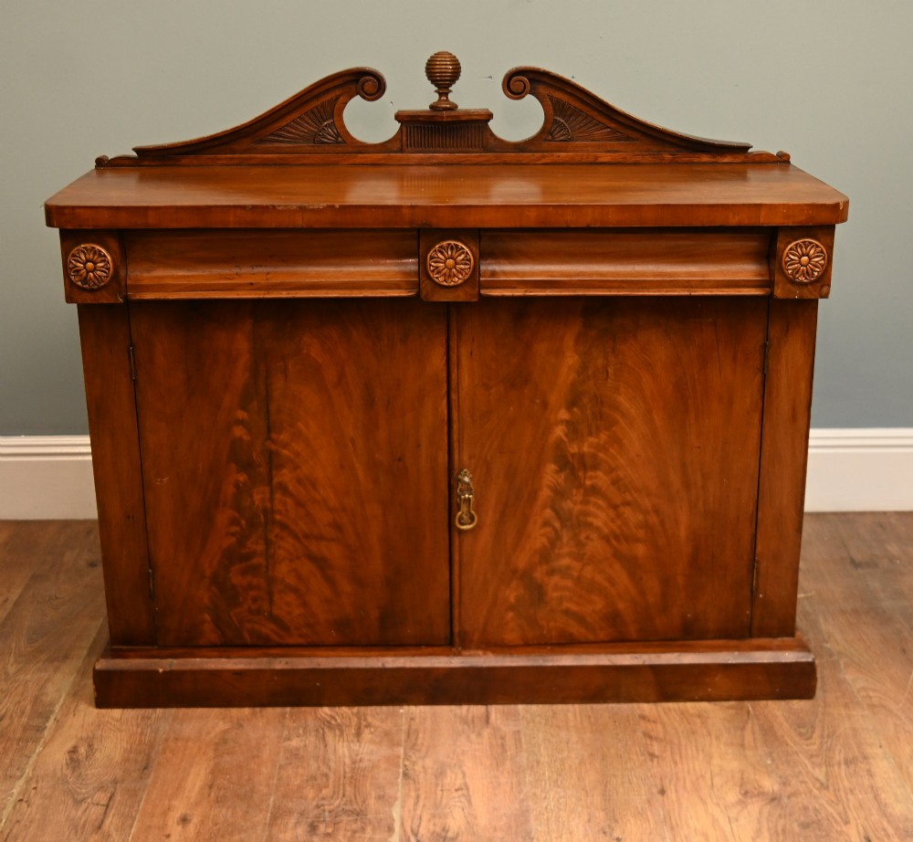 A William IV mahogany chiffonier side cabinet featuring a scroll-carved back gallery, central turned finial, carved rosette accents, and twin flame mahogany cupboard doors.