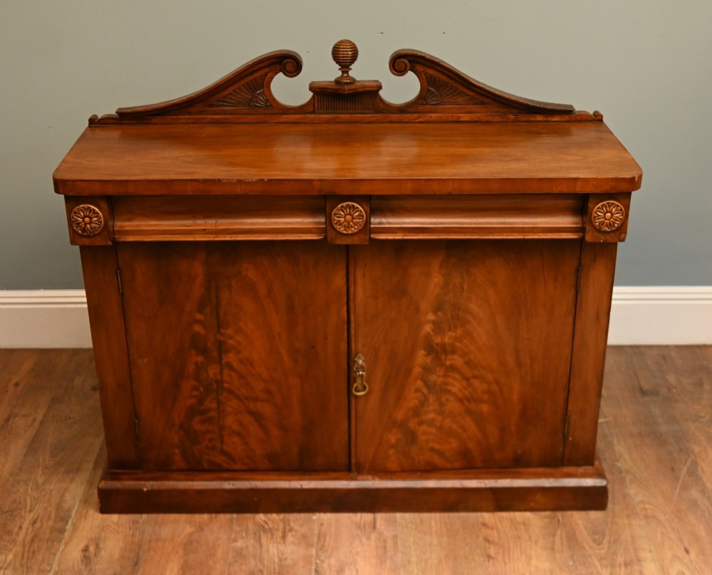 A William IV mahogany chiffonier side cabinet featuring a scroll-carved back gallery, central turned finial, carved rosette accents, and twin flame mahogany cupboard doors. View II