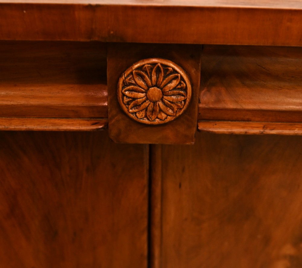 A William IV mahogany chiffonier side cabinet featuring a scroll-carved back gallery, central turned finial, carved rosette accents, and twin flame mahogany cupboard doors. View V