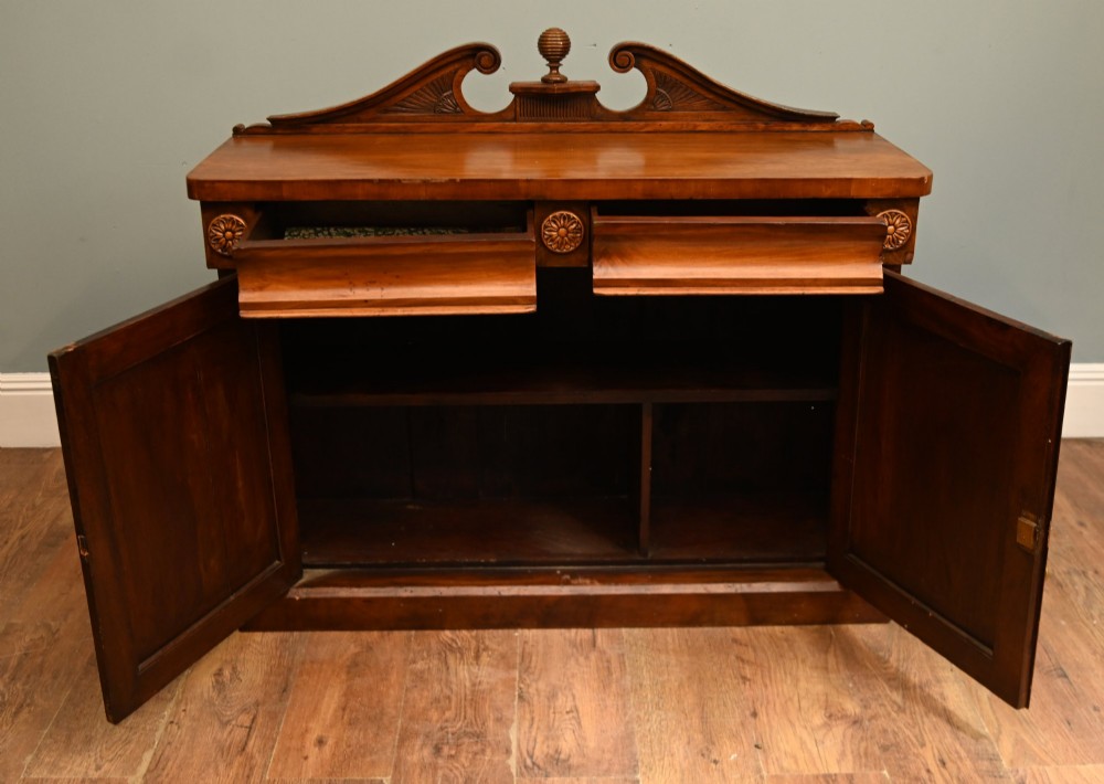A William IV mahogany chiffonier side cabinet featuring a scroll-carved back gallery, central turned finial, carved rosette accents, and twin flame mahogany cupboard doors. View VI