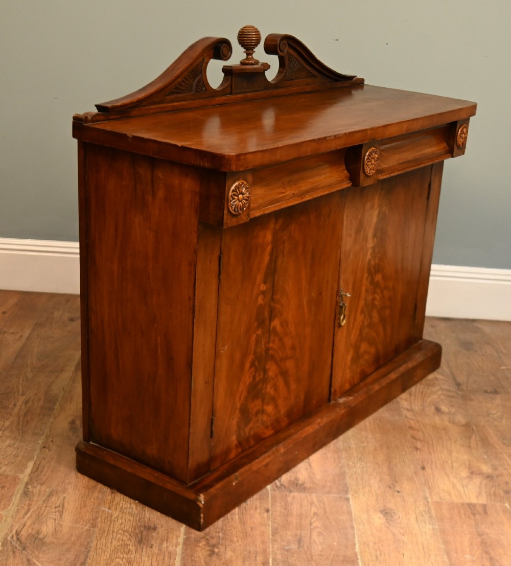 A William IV mahogany chiffonier side cabinet featuring a scroll-carved back gallery, central turned finial, carved rosette accents, and twin flame mahogany cupboard doors. View VII