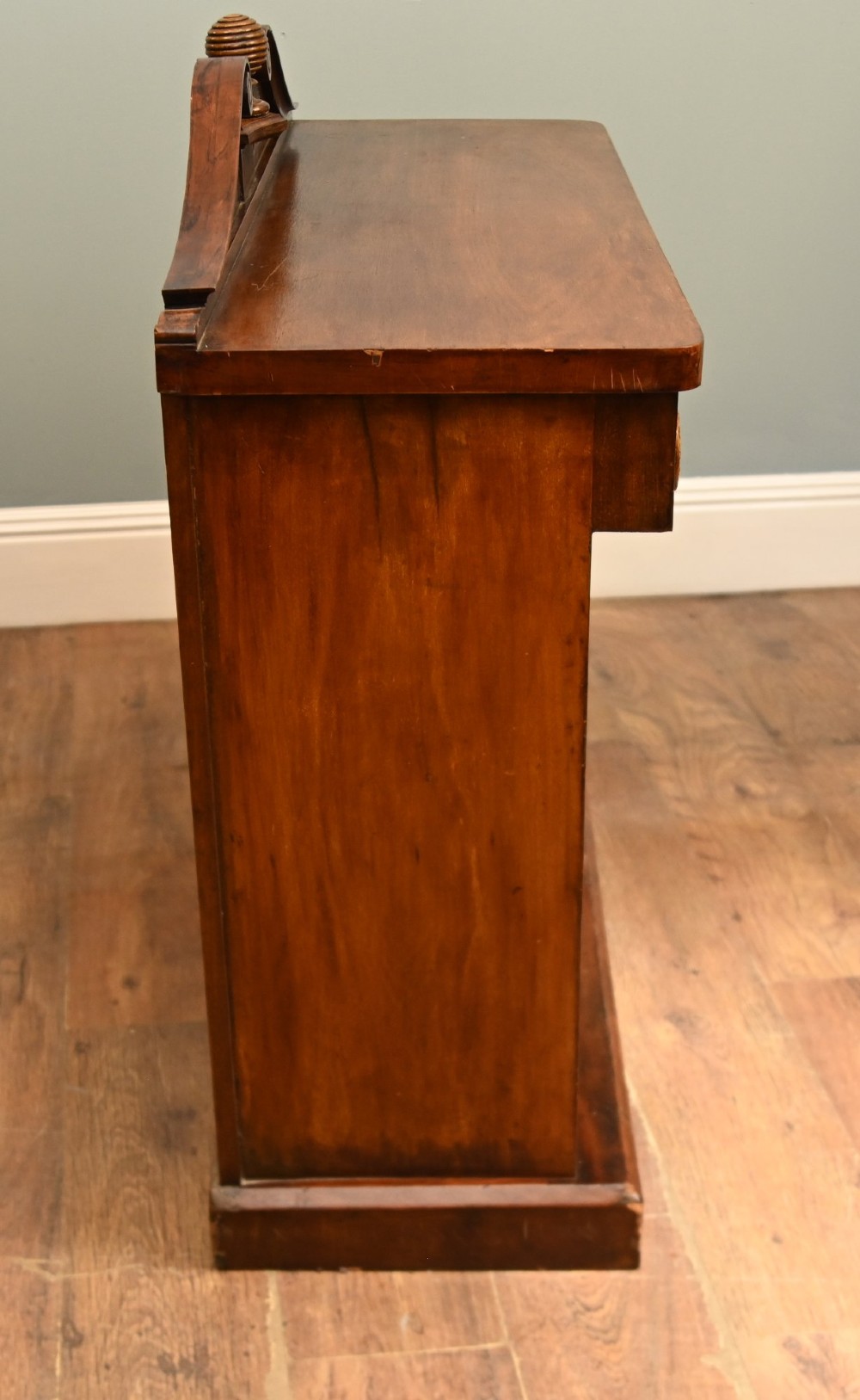 A William IV mahogany chiffonier side cabinet featuring a scroll-carved back gallery, central turned finial, carved rosette accents, and twin flame mahogany cupboard doors. View IX