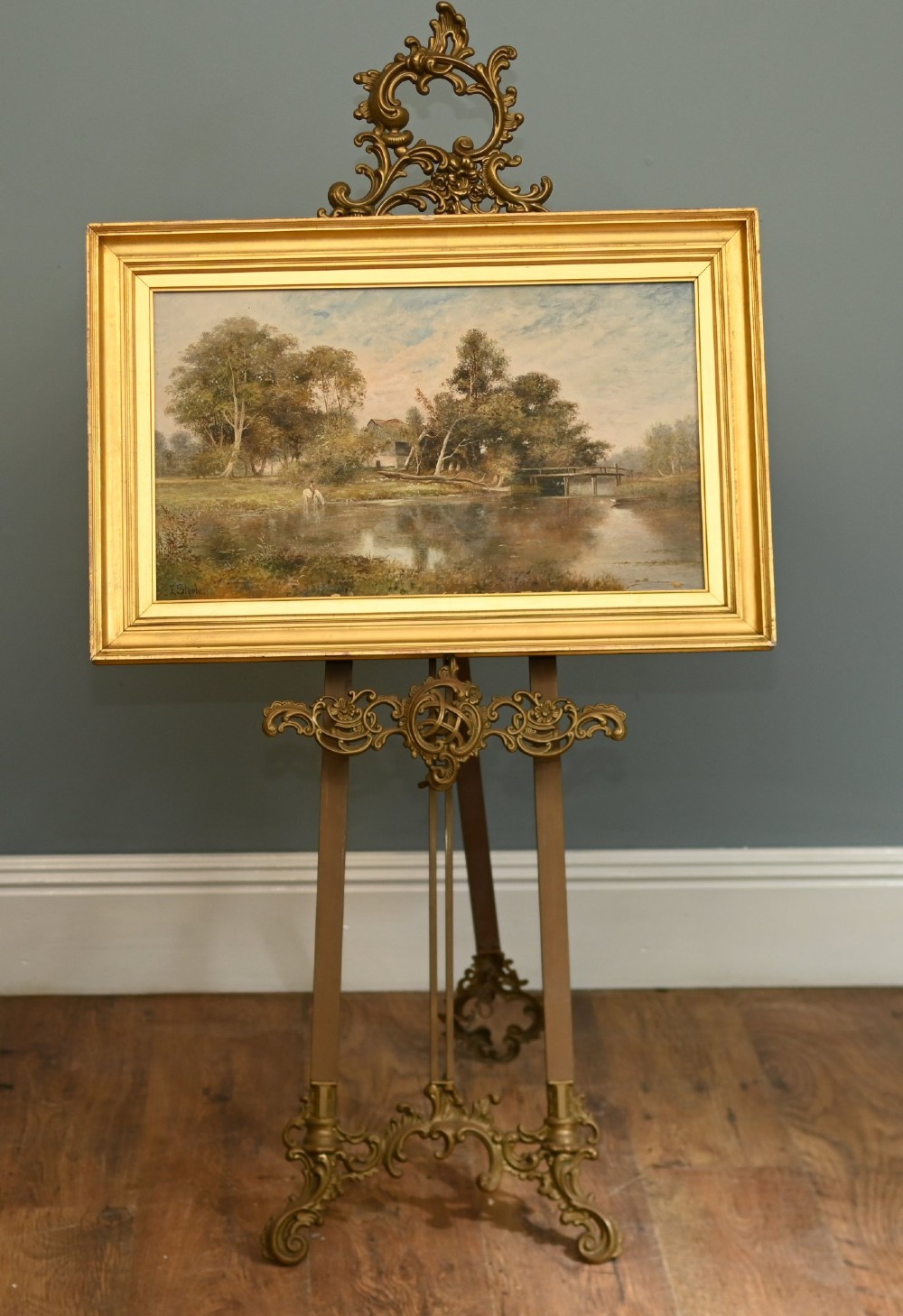 A 19th-century Victorian oil painting depicting a river landscape with trees and a wooden bridge, housed in a moulded gold gilt frame.