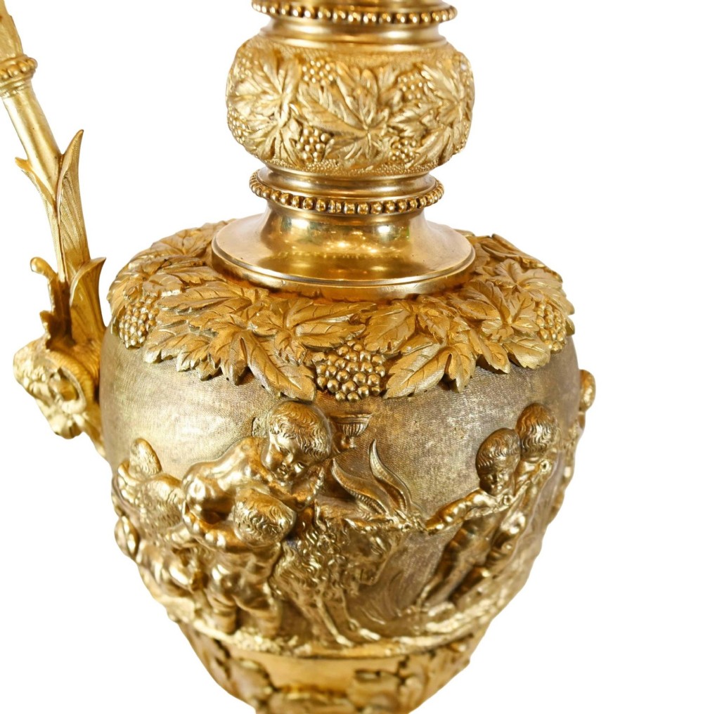 A pair of 19th-century French gilt bronze ewers featuring detailed Bacchus relief scenes, leaf motifs, and cherub-topped handles. View V