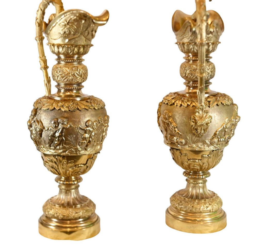 A pair of 19th-century French gilt bronze ewers featuring detailed Bacchus relief scenes, leaf motifs, and cherub-topped handles. View X