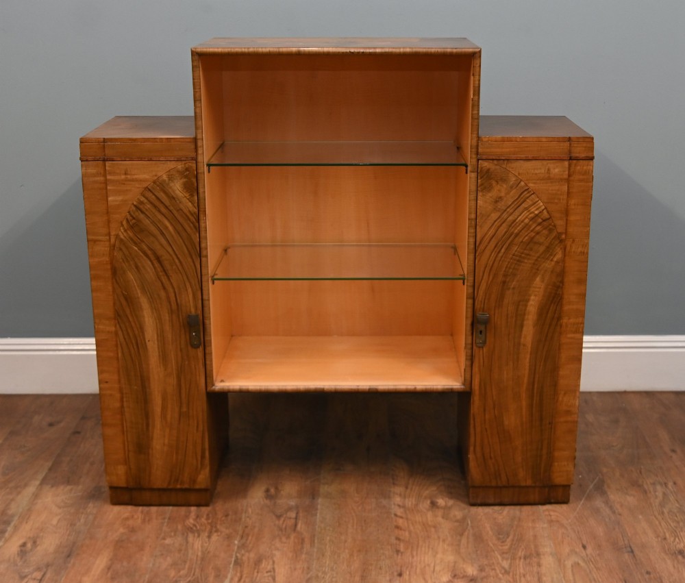 An Art Deco figured walnut display cabinet with a raised central open section containing two glass shelves, flanked by two side cupboards with arched veneer detailing.