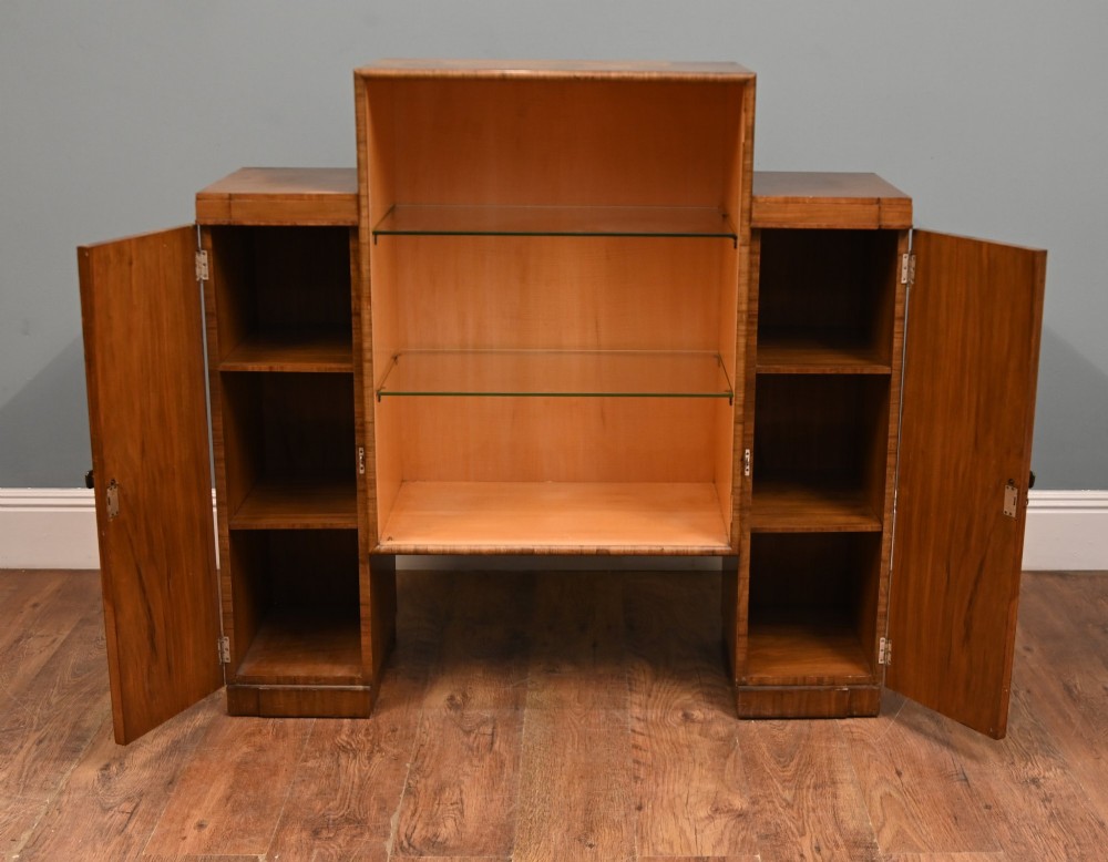 An Art Deco figured walnut display cabinet with a raised central open section containing two glass shelves, flanked by two side cupboards with arched veneer detailing. View II