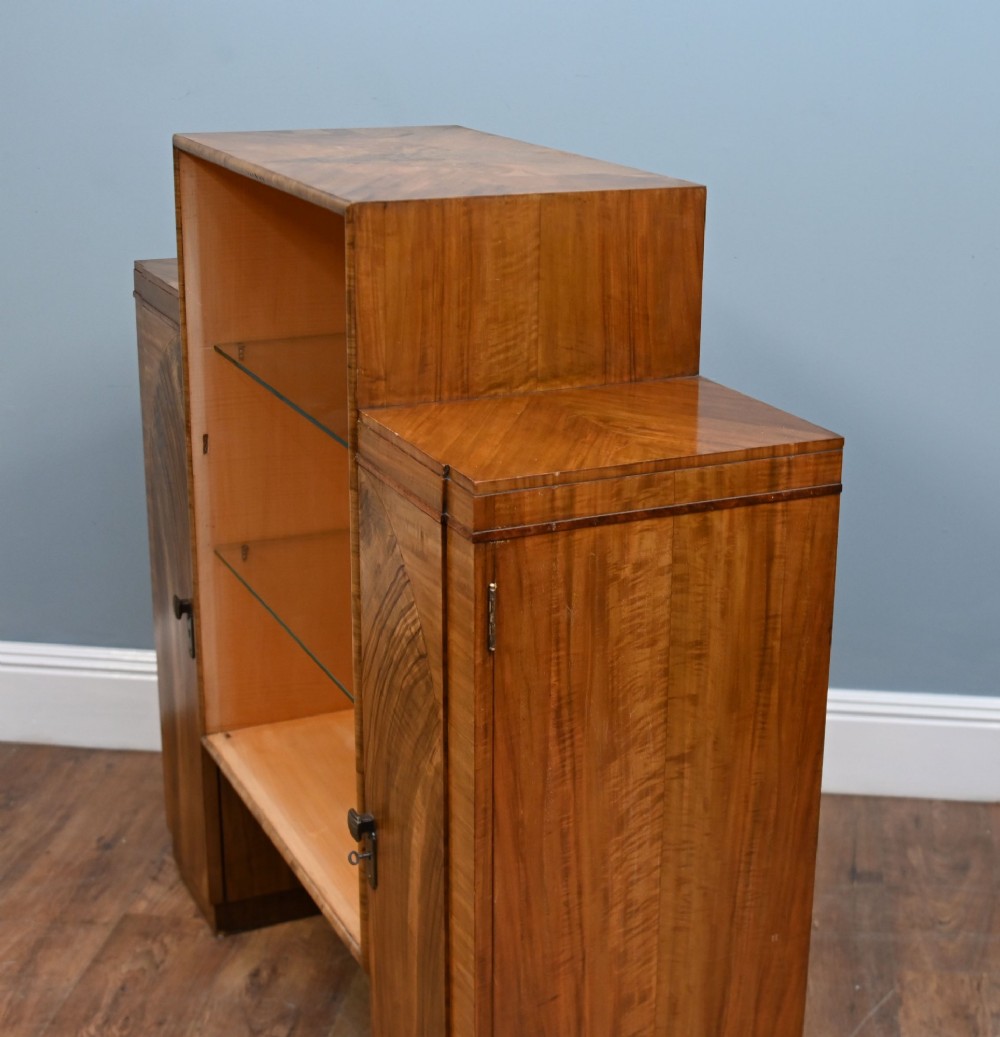 An Art Deco figured walnut display cabinet with a raised central open section containing two glass shelves, flanked by two side cupboards with arched veneer detailing. View V
