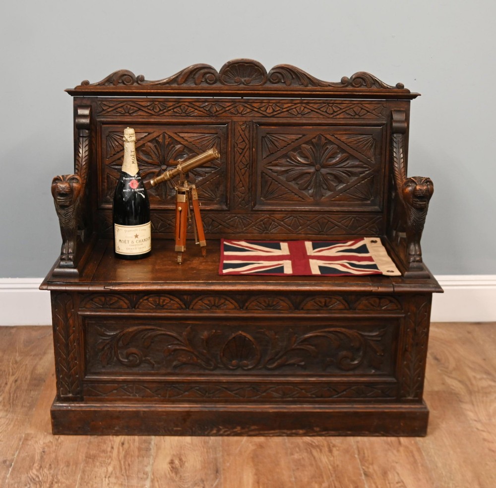 A Victorian dark oak carved settle featuring carved lion head armrests, detailed floral and geometric back panels, and a lift-up storage seat. View II