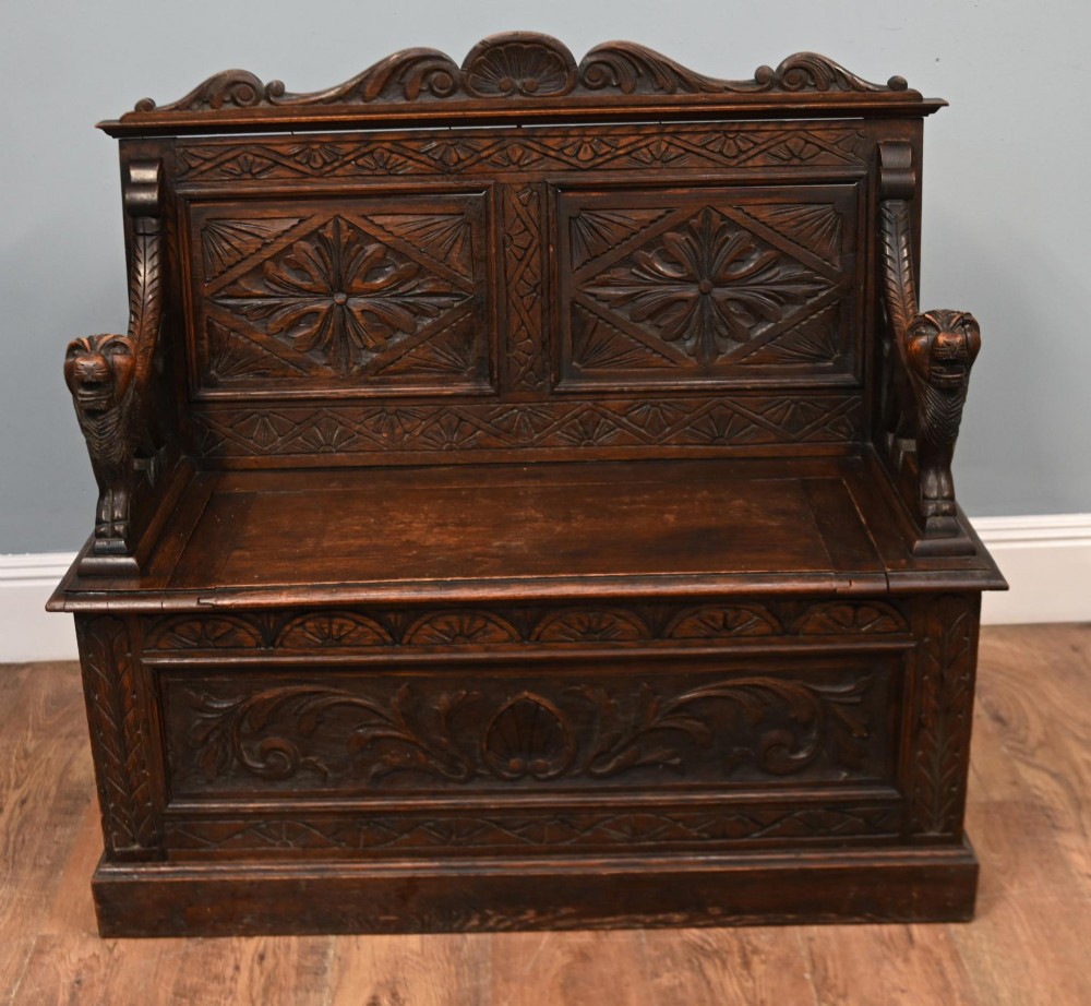 A Victorian dark oak carved settle featuring carved lion head armrests, detailed floral and geometric back panels, and a lift-up storage seat.
