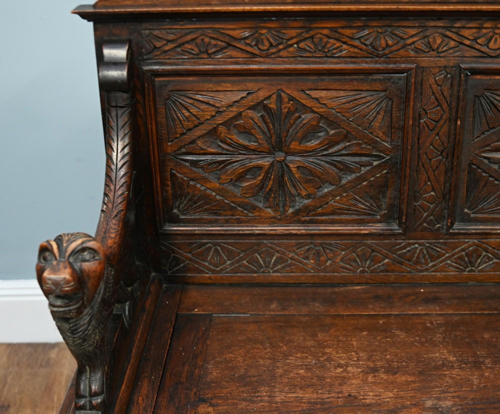 A Victorian dark oak carved settle featuring carved lion head armrests, detailed floral and geometric back panels, and a lift-up storage seat. View IV