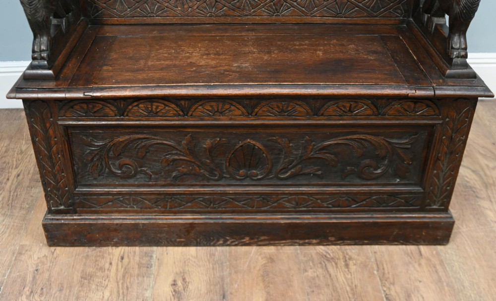 A Victorian dark oak carved settle featuring carved lion head armrests, detailed floral and geometric back panels, and a lift-up storage seat. View V