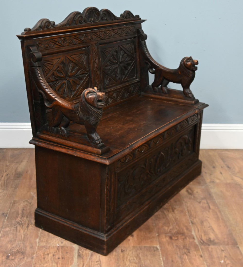 A Victorian dark oak carved settle featuring carved lion head armrests, detailed floral and geometric back panels, and a lift-up storage seat. View VI