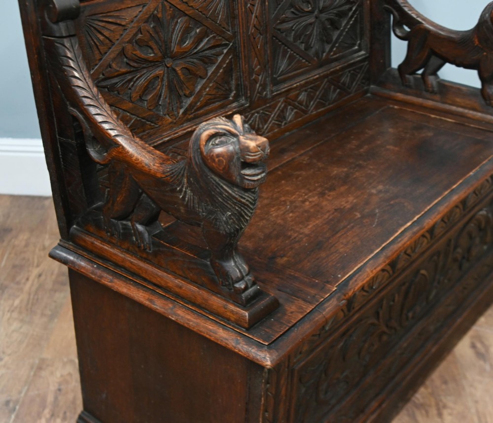A Victorian dark oak carved settle featuring carved lion head armrests, detailed floral and geometric back panels, and a lift-up storage seat. View VII
