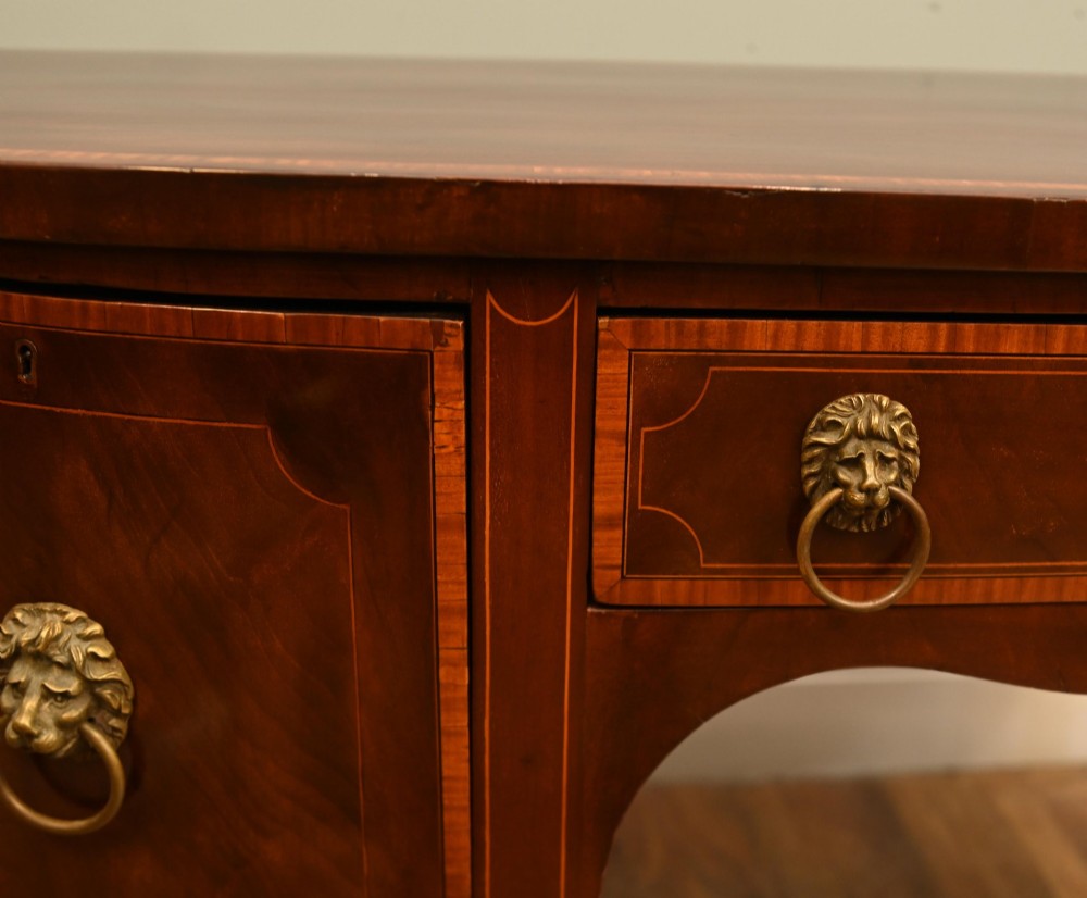 An Edwardian mahogany bowfront sideboard with satinwood inlaid borders, three central drawer and cabinet sections, and brass lion mask ring handles on tapered legs. View VI