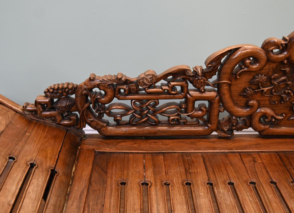 An intricately hand-carved warm brown hardwood Chinese opium bench featuring a slatted seat, elevated recline end, and pierced scrollwork along the backrest. View V