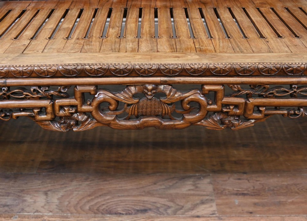 An intricately hand-carved warm brown hardwood Chinese opium bench featuring a slatted seat, elevated recline end, and pierced scrollwork along the backrest. View VI