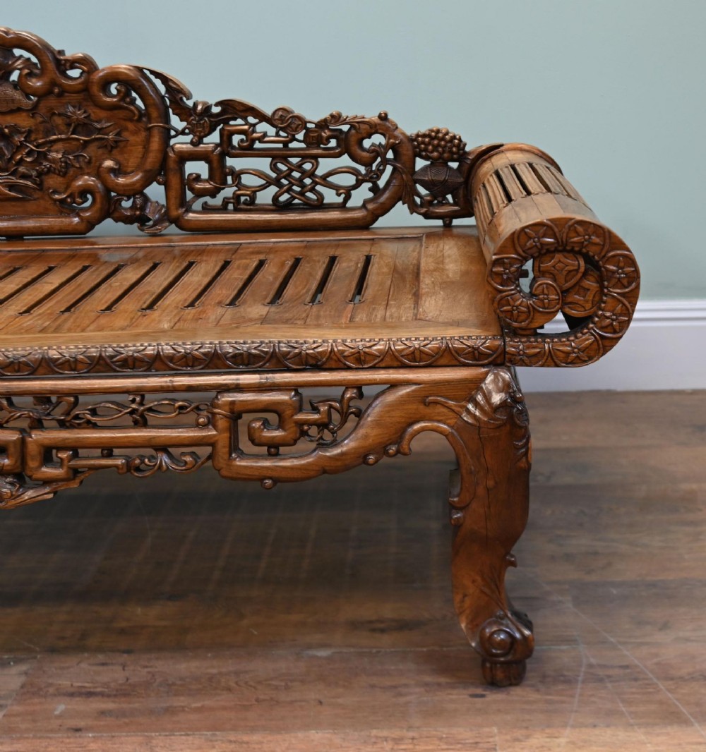 An intricately hand-carved warm brown hardwood Chinese opium bench featuring a slatted seat, elevated recline end, and pierced scrollwork along the backrest. View VII