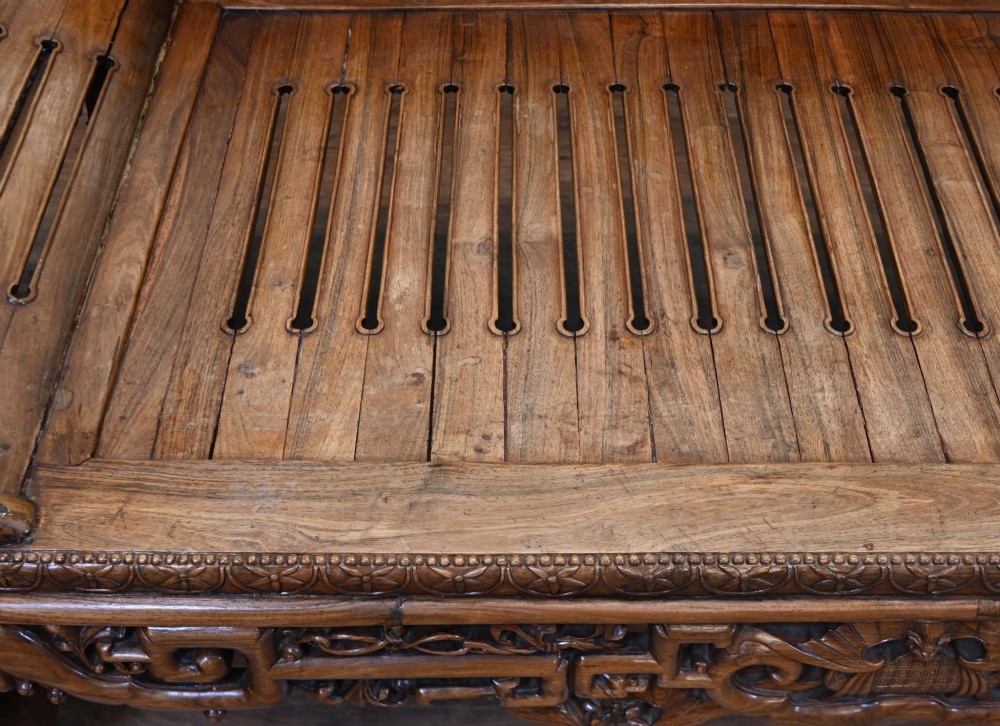An intricately hand-carved warm brown hardwood Chinese opium bench featuring a slatted seat, elevated recline end, and pierced scrollwork along the backrest. View VIII
