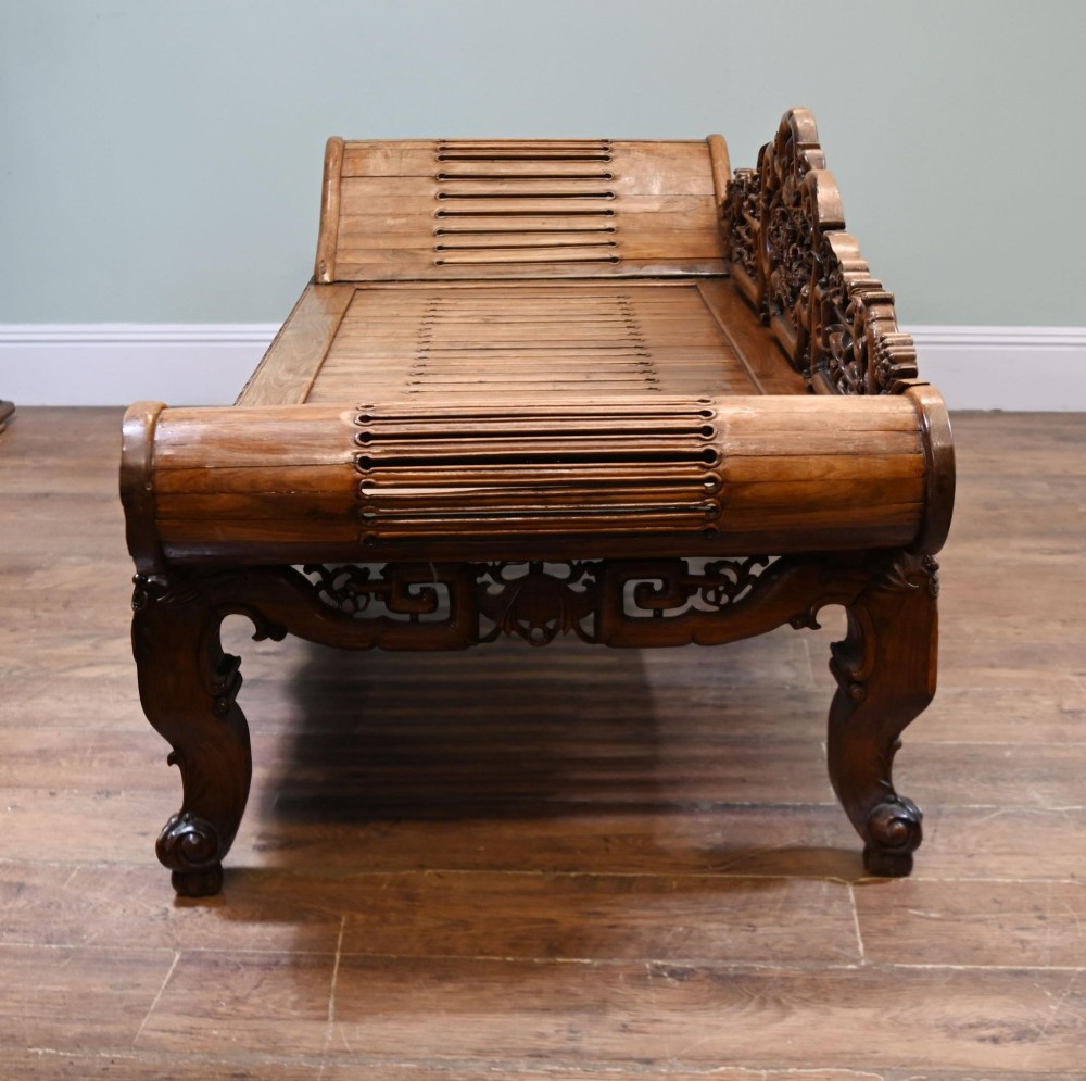 An intricately hand-carved warm brown hardwood Chinese opium bench featuring a slatted seat, elevated recline end, and pierced scrollwork along the backrest. View IX