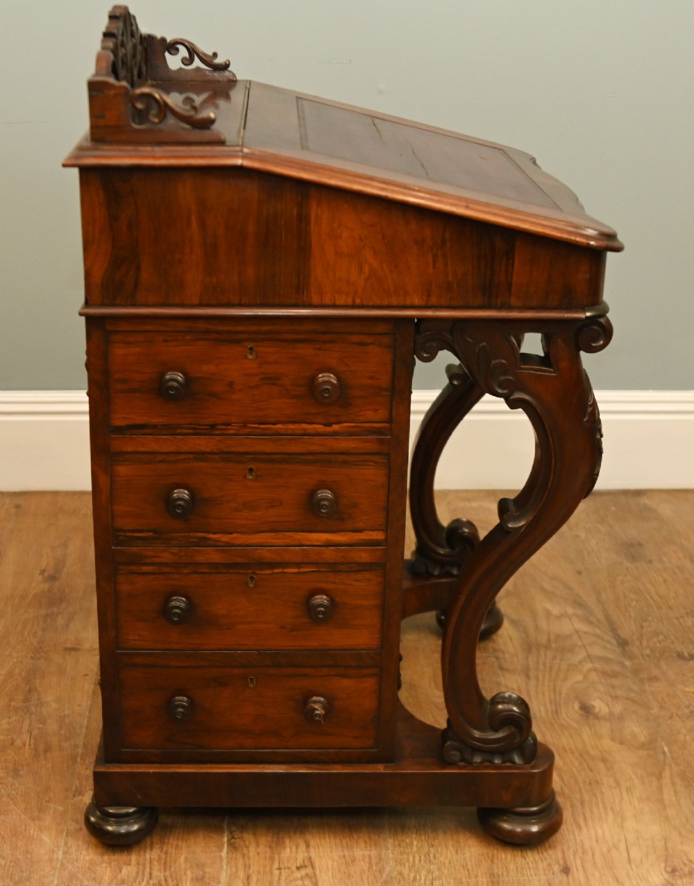 A Victorian mahogany Davenport desk featuring a sloped leather writing top, carved cresting rail, serpentine front supports, and side drawers. View X