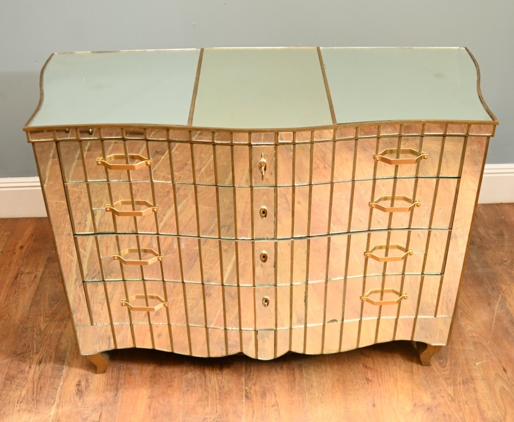 An Art Deco mirrored chest of drawers featuring segmented glass panelling, four drawers with geometric brass handles, and a shaped lower apron.