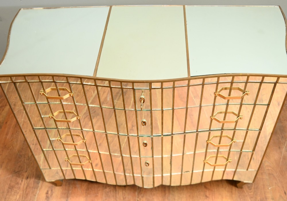 An Art Deco mirrored chest of drawers featuring segmented glass panelling, four drawers with geometric brass handles, and a shaped lower apron. View V