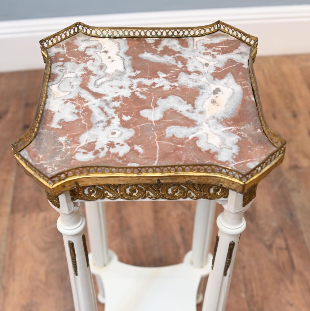 A white painted tall French pedestal stand featuring four fluted legs, gilt brass mounts, a lower tier, and a shaped variegated marble top with a brass gallery. View IV
