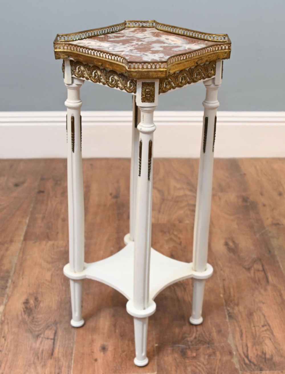 A white painted tall French pedestal stand featuring four fluted legs, gilt brass mounts, a lower tier, and a shaped variegated marble top with a brass gallery.