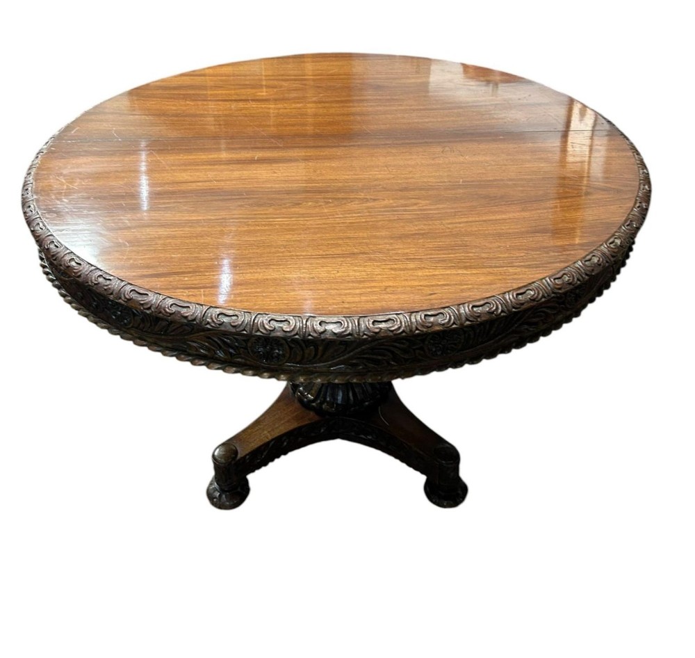 A round antique Burmese hardwood dining table featuring a polished top, an intricately carved apron, and a central pedestal base with tripod legs.