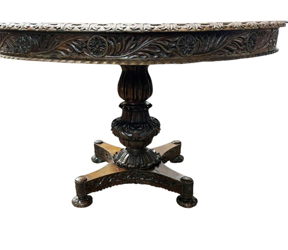 A round antique Burmese hardwood dining table featuring a polished top, an intricately carved apron, and a central pedestal base with tripod legs. View III