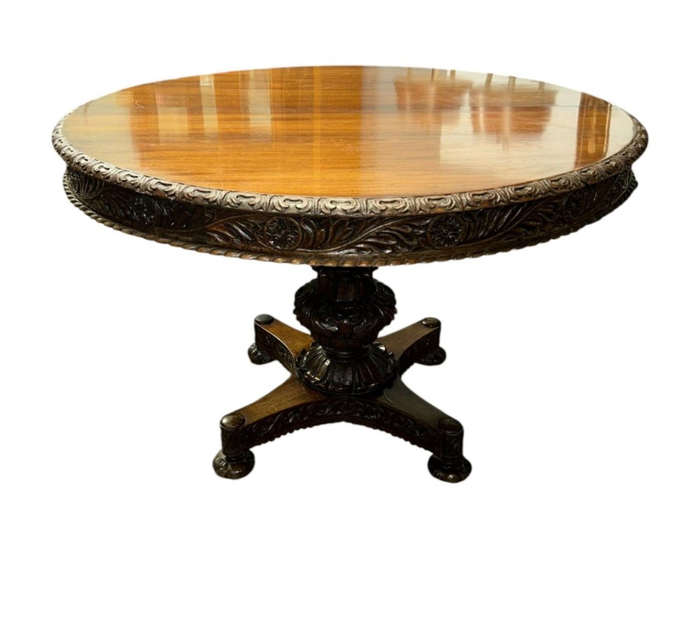 A round antique Burmese hardwood dining table featuring a polished top, an intricately carved apron, and a central pedestal base with tripod legs. View IV