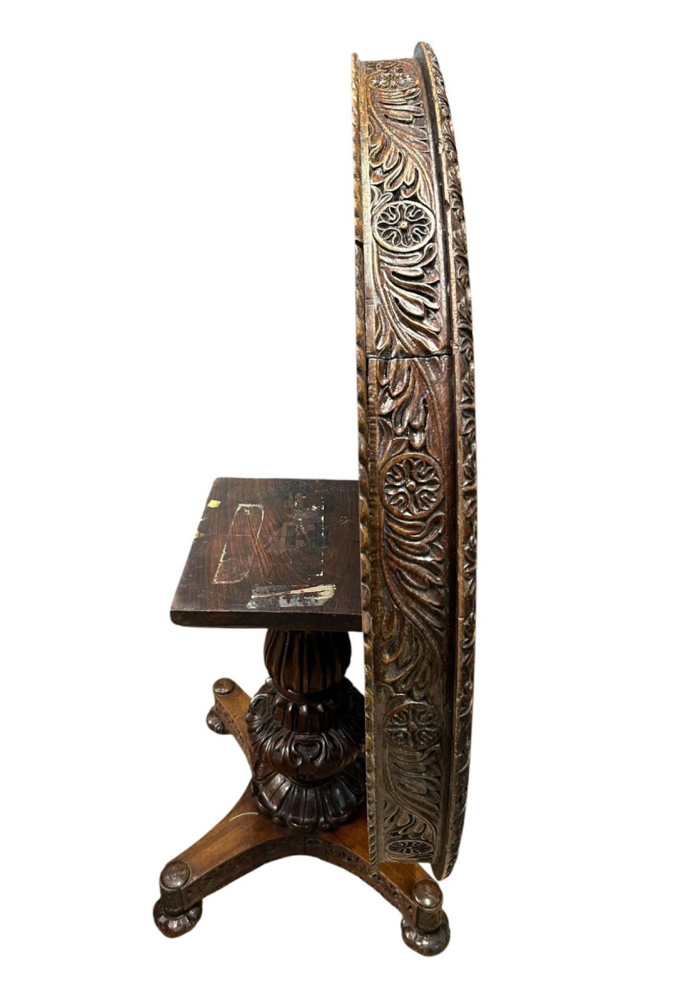 A round antique Burmese hardwood dining table featuring a polished top, an intricately carved apron, and a central pedestal base with tripod legs. View VI