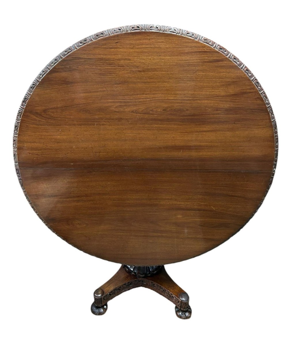 A round antique Burmese hardwood dining table featuring a polished top, an intricately carved apron, and a central pedestal base with tripod legs. View IX
