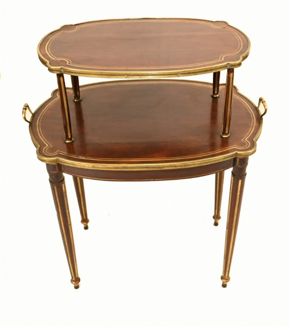 A two-tiered mahogany side table featuring shaped oval tops edged with brass galleries, slender turned legs, and brass side handles on the lower tier.