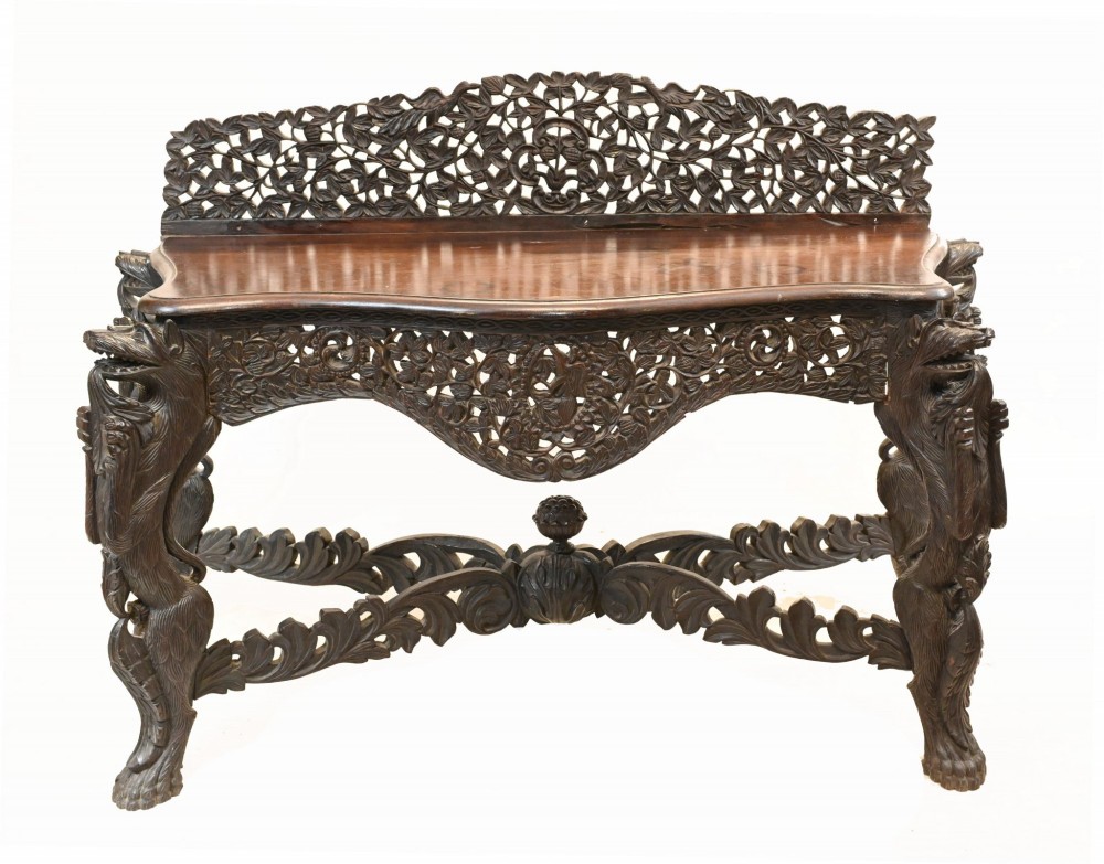A late 19th-century Burmese dark teak console table featuring intricately pierced foliate carvings on the gallery back, skirt, and cross-stretcher, supported by legs carved as mythical figures.