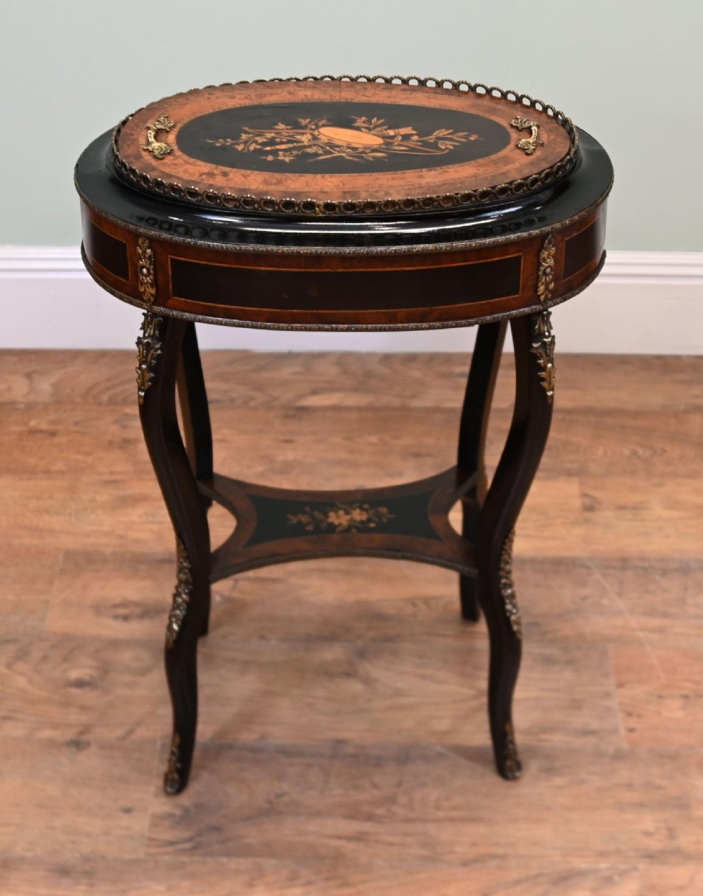 An antique French oval jardiniere table featuring amboyna wood veneer, floral marquetry, gilt-metal mounts, and ebonised cabriole legs.