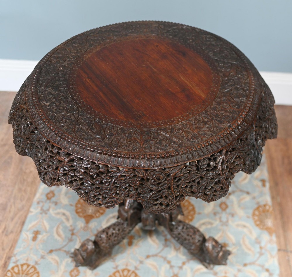 An antique dark hardwood Burmese side table with intricate pierced foliage carving around the circular top and a carved tripod base featuring mythological figure feet. View V