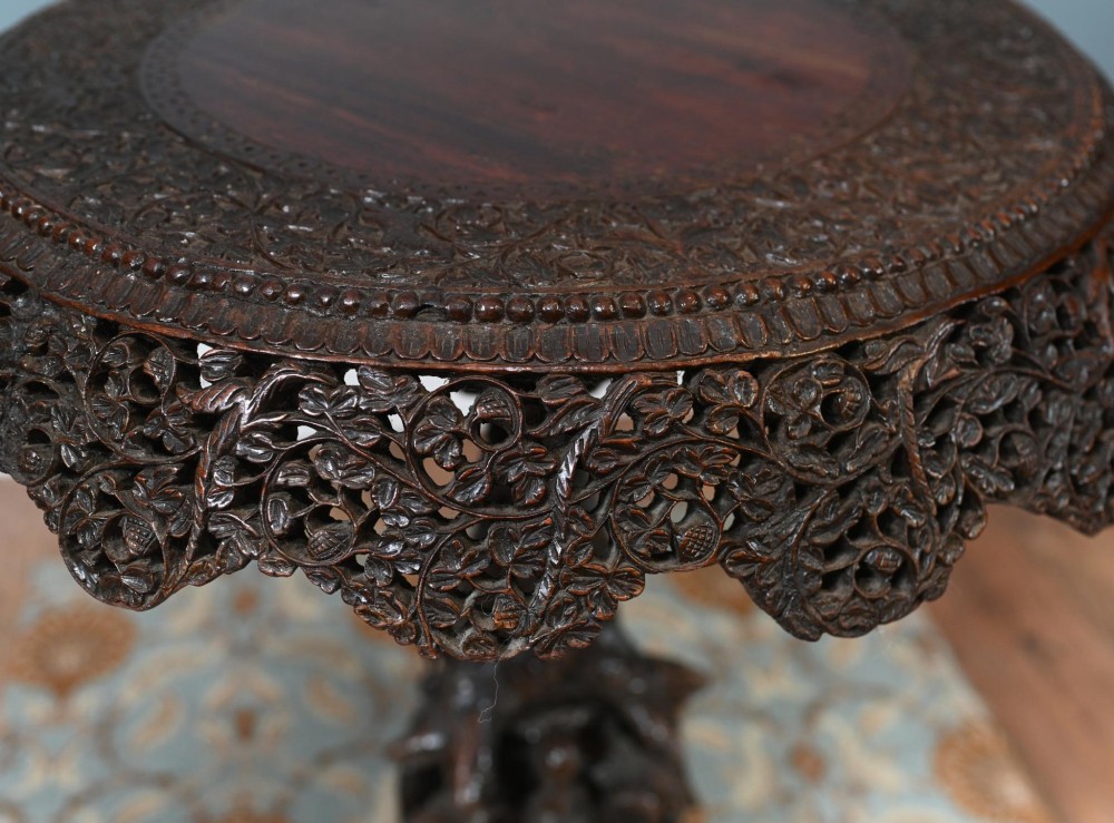 An antique dark hardwood Burmese side table with intricate pierced foliage carving around the circular top and a carved tripod base featuring mythological figure feet. View VI