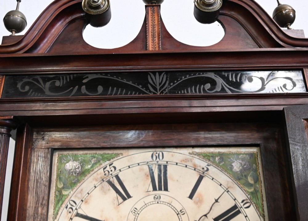 A Georgian mahogany longcase clock featuring a swan-neck pediment with three brass finials, a painted dial with Roman numerals, and a flame mahogany door. View IV