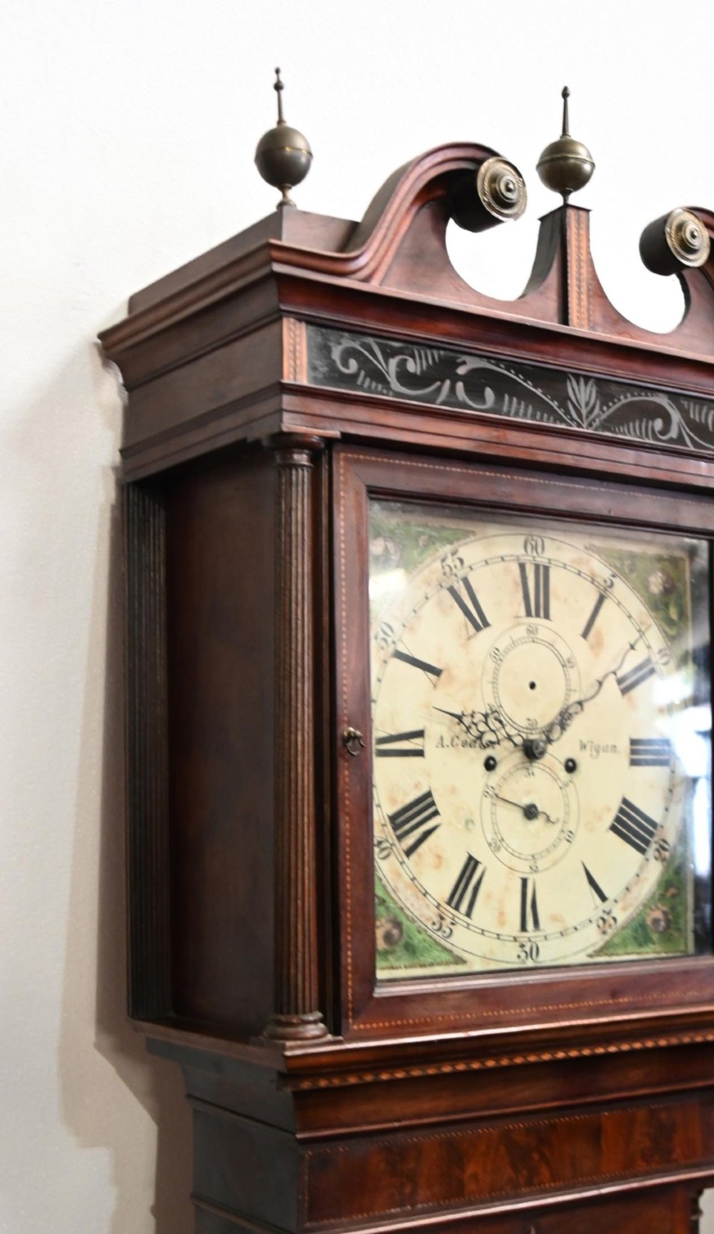 A Georgian mahogany longcase clock featuring a swan-neck pediment with three brass finials, a painted dial with Roman numerals, and a flame mahogany door. View VI