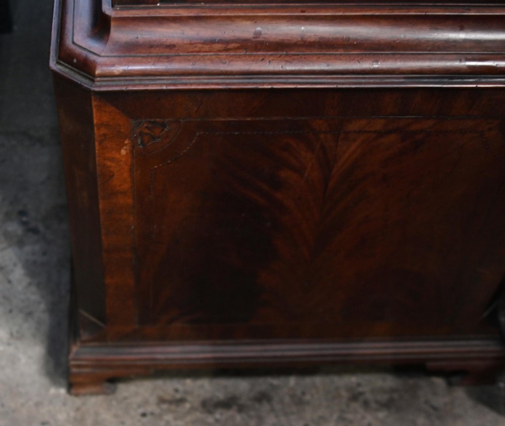 A Georgian mahogany longcase clock featuring a swan-neck pediment with three brass finials, a painted dial with Roman numerals, and a flame mahogany door. View IX