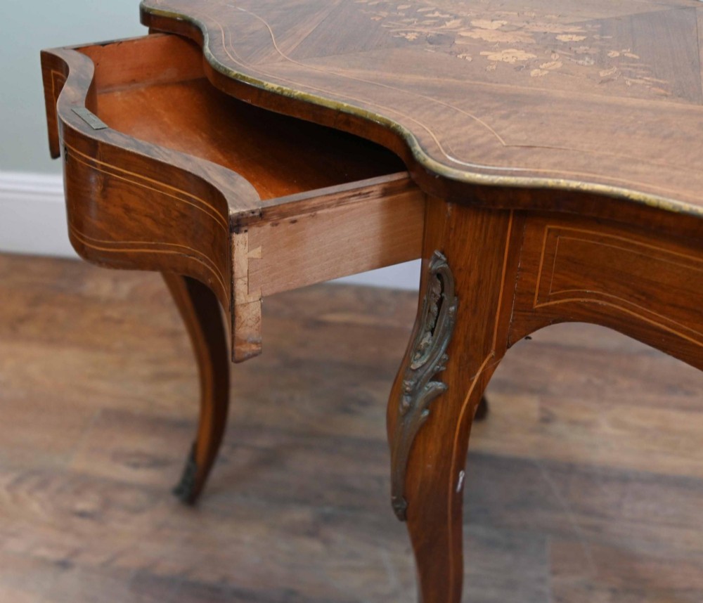 A French Louis XV style writing desk crafted from brown wood with a serpentine top, floral marquetry inlay, cabriole legs, and brass mounts. View IX