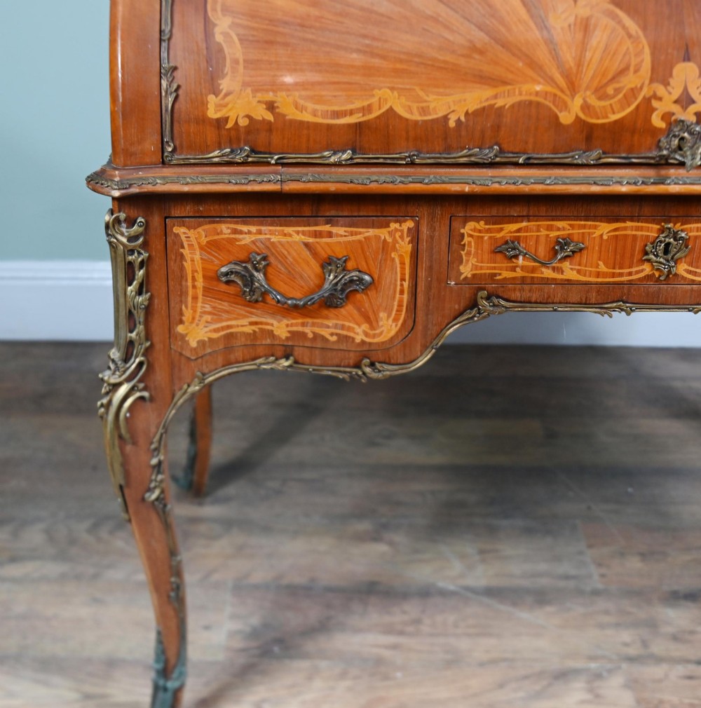 An antique French roll-top bureau featuring wood marquetry inlay, decorative brass mounts, cabriole legs, and drawers. View V