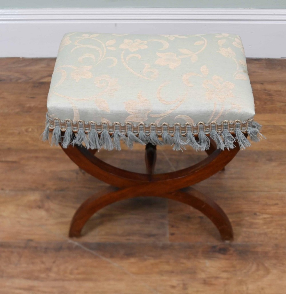 A Regency style mahogany stool featuring curved x-frame legs, a central turned stretcher, and a pale blue patterned damask upholstered seat finished with grey fringe trim. View VI
