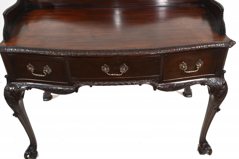 A dark mahogany Victorian console table featuring three drawers with decorative brass handles, carved cabriole legs, and a low gallery back. View VI