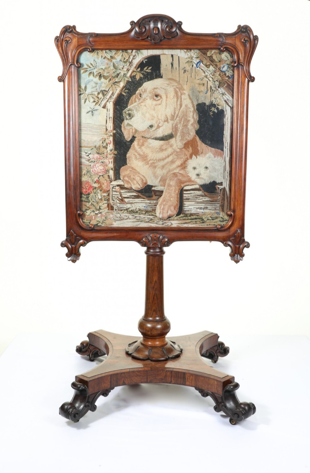 A Victorian carved mahogany fire screen featuring a needlepoint tapestry panel depicting a large hound and a small terrier inside a wooden kennel.